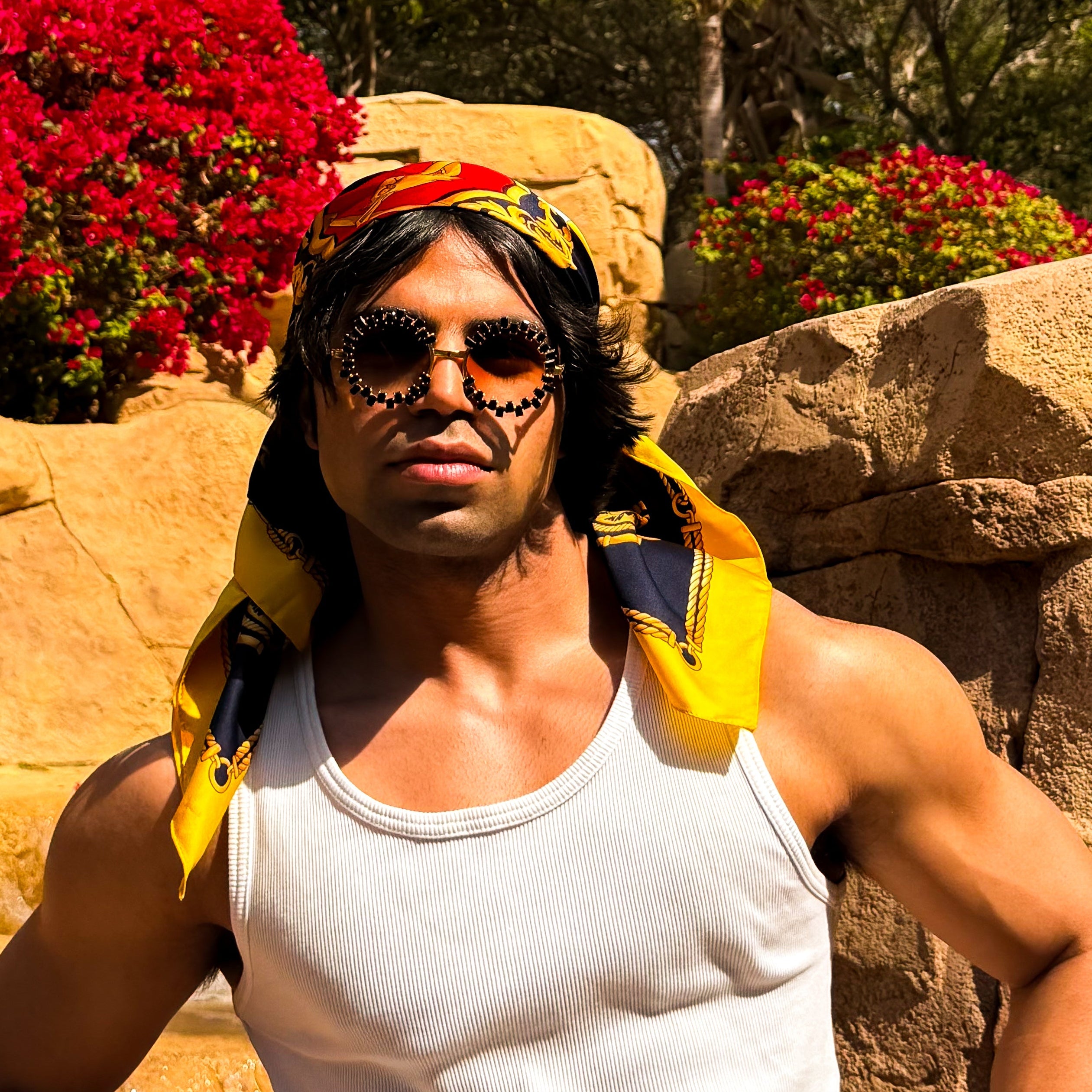 Person wearing a white tank top and yellow jacket with sunglasses, standing against a natural background with rocks and flowers.